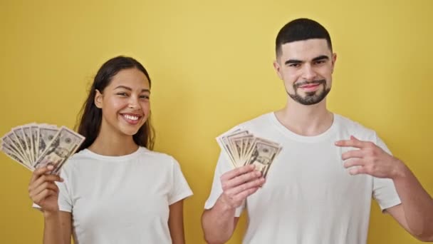 Zelfverzekerd mooi koppel glimlachend en wijzend naar dollars, het leven van de gelukkige cash lifestyle samen tegen een levendige gele geïsoleerde achtergrond - Video