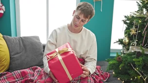 Νεαρός καυκάσιος άνδρας αγκαλιάζει χριστουγεννιάτικο δώρο κάθεται στον καναπέ στο σπίτι - Πλάνα, βίντεο