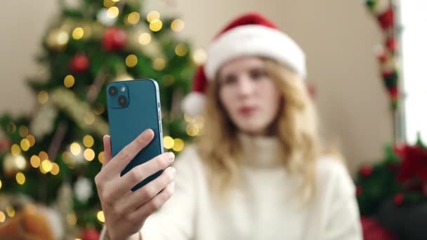 Young blonde woman having video call sitting on sofa by christmas tree at home - Кадры, видео