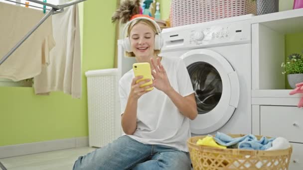 Młoda blondynka korzystająca ze smartfona i słuchawek uśmiechających się do pralni - Materiał filmowy, wideo