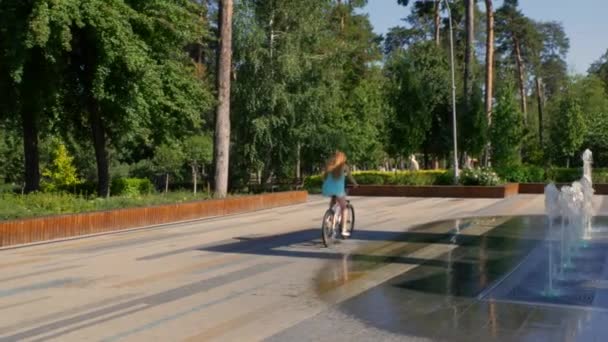 Прогулянка міським парком на велосипеді. Дівчина їде вздовж алеї біля фонтану, навколо ялинки та соснових дерев. Високоякісні 4k кадри - Кадри, відео