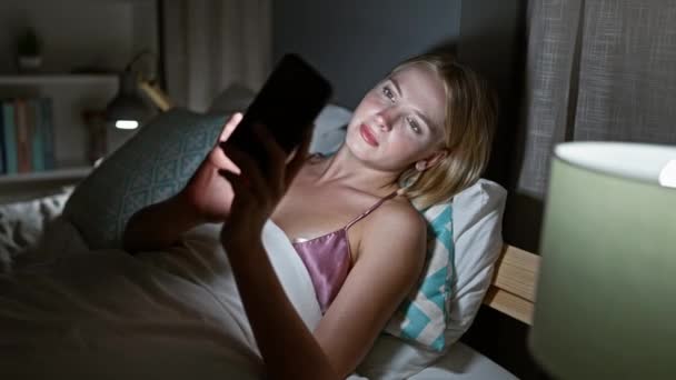 Fiatal szőke nő használ smartphone feküdt az ágyon mosolyog a hálószobában - Felvétel, videó