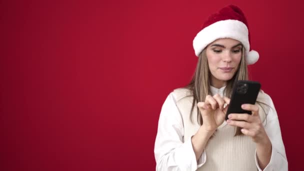 Młoda, piękna Latynoska kobieta w świątecznym kapeluszu za pomocą smartfona na izolowanym czerwonym tle - Materiał filmowy, wideo
