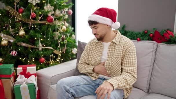 Hombre árabe joven sufriendo dolor de estómago sentado junto al árbol de Navidad en casa - Metraje, vídeo