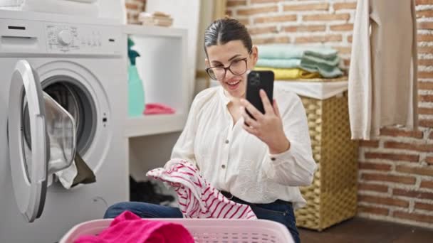 Młoda piękna Latynoska robi pranie wykonując wideo połączenie ze smartfonem w pralni - Materiał filmowy, wideo
