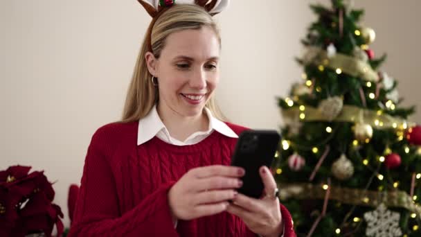 Mujer rubia joven hacer selfie por teléfono inteligente de pie junto al árbol de Navidad en casa - Metraje, vídeo