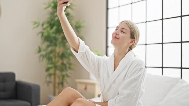 Fiatal szőke nő visel fürdőköpeny ül az ágyban, hogy szelfi okostelefon a hálószobában - Felvétel, videó