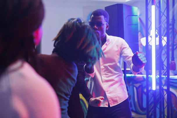 Cooler afrikanisch-amerikanischer Mann feiert und tanzt in einem Nachtclub, der mit leuchtenden Lichtern beleuchtet ist. Clubber haben Spaß auf der Tanzfläche in einem überfüllten Club, während sie Diskothek besuchen - Foto, Bild