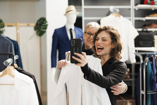 Playful stylish women taking picture with modern phone in showroom, having fun during shopping session. Cheerful clients checking casual wear brands for commercial activity, buying trendy clothes - Fotoğraf, Görsel