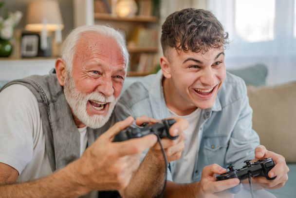 Caucasian teenager and senior man grandfather and grandson sit on sofa bed at home play console video game hold joystick controller have fun family bonding males men - Photo, Image