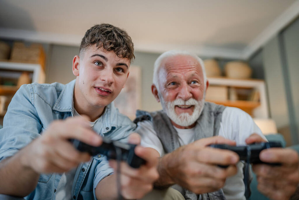 コーカサスティーンエイジャーとシニア男祖父と孫は,ホームプレイコンソールビデオゲームホールジョイスティックコントローラーでソファーベッドに座って楽しい家族の絆男性を持っています - 写真・画像