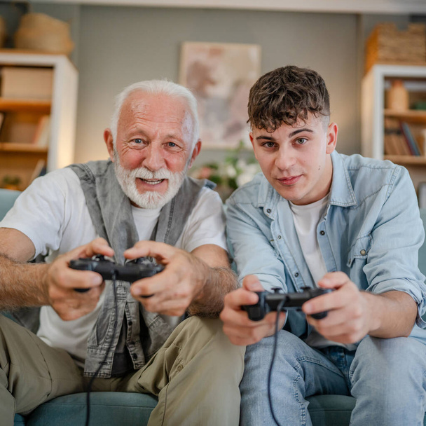 Caucasian teenager and senior man grandfather and grandson sit on sofa bed at home play console video game hold joystick controller have fun family bonding males men - Photo, Image