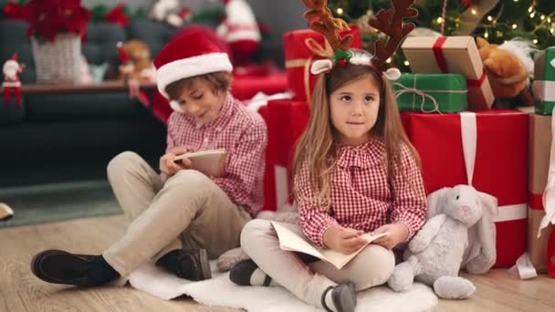 Αδελφός και αδελφή σχέδιο στο σημειωματάριο κάθεται στο πάτωμα από χριστουγεννιάτικο δέντρο στο σπίτι - Πλάνα, βίντεο
