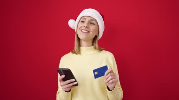 Mujer rubia joven haciendo compras en línea de Navidad con teléfono inteligente sobre fondo rojo aislado - Imágenes, Vídeo