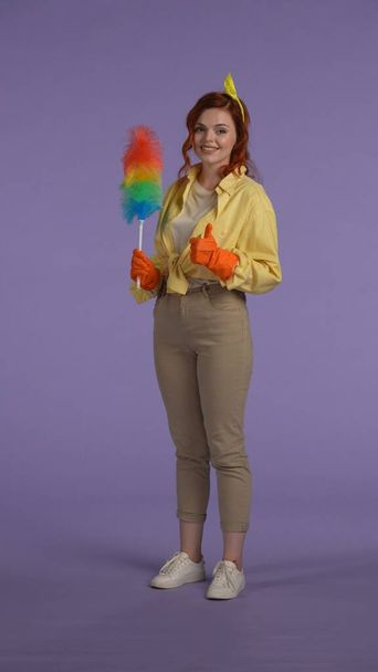 Everyday housekeeping creative concept. Vertical photo. Woman in casual clothing and rubber gloves holding pipiduster, showing thumbs up at the camera, smiling face expression. - Фото, зображення