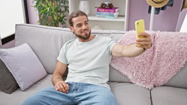 Giovane uomo ispanico scattare foto selfie con smartphone seduto sul divano a casa - Filmati, video