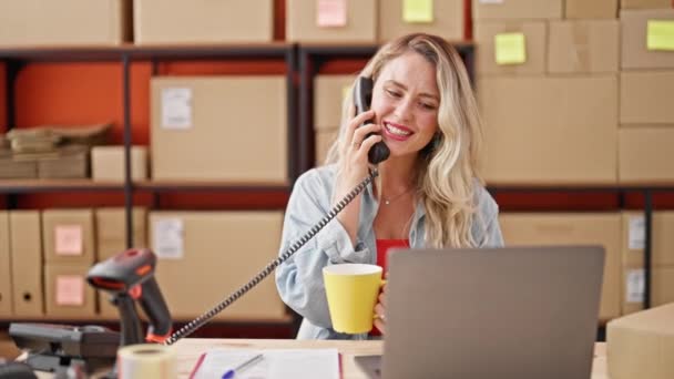 Νεαρή ξανθιά υπάλληλος ηλεκτρονικού εμπορίου που μιλάει στο τηλέφωνο πίνοντας καφέ στο γραφείο - Πλάνα, βίντεο