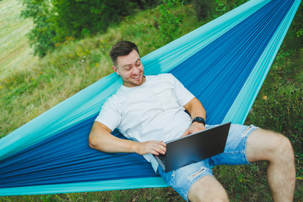 Egy fiatal férfi laptoppal egy függőágyban ül a természetben, és távolról dolgozik. Munka a természetben a nyaralás alatt. - Fotó, kép