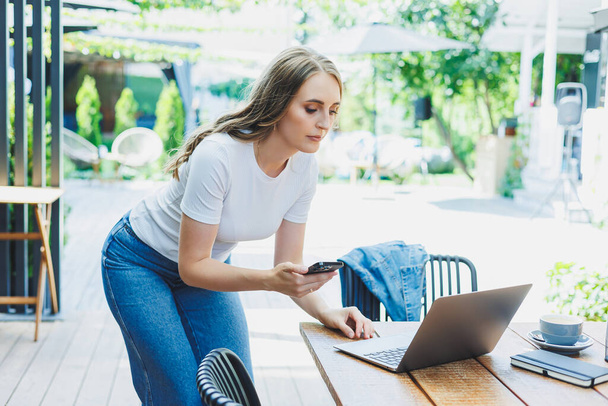 Modern bir kafe caddesindeki bir kafede terasta oturan genç bir kadın. Telefonla masada oturan ve dışarıda dizüstü bilgisayar kullanan bir kadın. Bir yazlık kafede uzaktan çalışma - Fotoğraf, Görsel