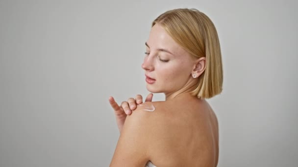 Mujer rubia joven aplicando crema en el hombro sobre fondo blanco aislado - Imágenes, Vídeo