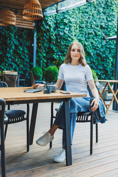 Eine nette Frau trinkt Kaffee auf der Terrasse eines Sommercafés und arbeitet an einem Laptop. Fernarbeit im Urlaub im Café mit Laptop. - Foto, Bild