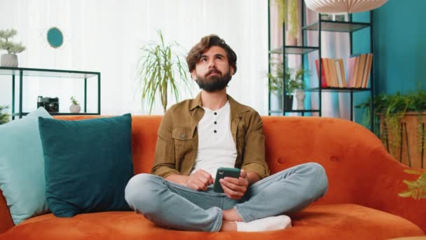 Middle eastern man sits on couch uses mobile phone smiles at home living room apartment. Young guy texting share messages content on smartphone social media applications online, watching relax movie - Footage, Video