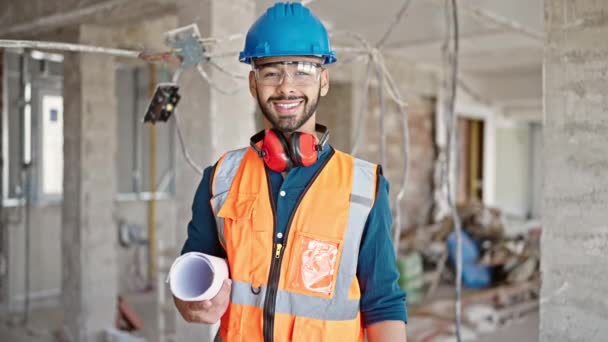 Jonge Spaanse man bouwer glimlachend vol vertrouwen staand met blauwdrukken op bouwplaats - Video