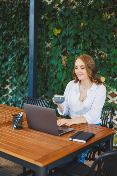 Young woman in casual clothes drinking coffee and talking on the phone, working on a green terrace at a laptop, looking at the screen and working on a project remotely from work in a cafe - Photo, Image