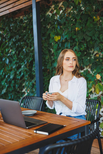 Egy fiatal nő alkalmi ruhában kávét iszik és telefonon beszél, egy zöld teraszon dolgozik egy laptopnál, nézi a képernyőt, és távolról dolgozik egy projekten egy kávézóban. - Fotó, kép