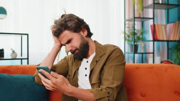 Surullinen tyytymätön mies käyttää älypuhelimen kirjoittamista selailu, menettää tulossa yllättynyt äkillinen arpajaiset pelin tuloksia, huonoja uutisia, onni menetys, epäonnistua, määräaika, virus. Nuori Lähi-idän kaveri kotona huoneessa sohvalla - Materiaali, video