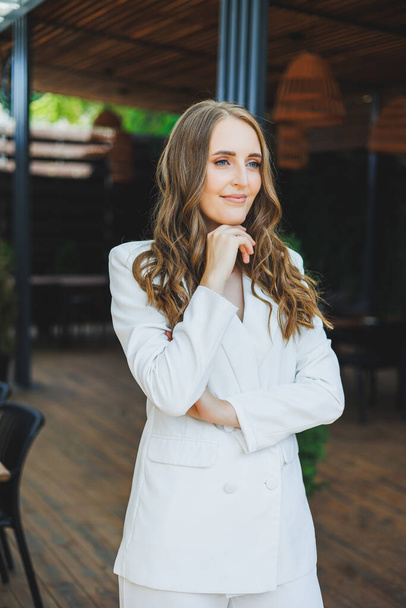 A young stylish woman in a white classic suit with a smile on her face is standing on the terrace of a summer cafe. - Photo, Image