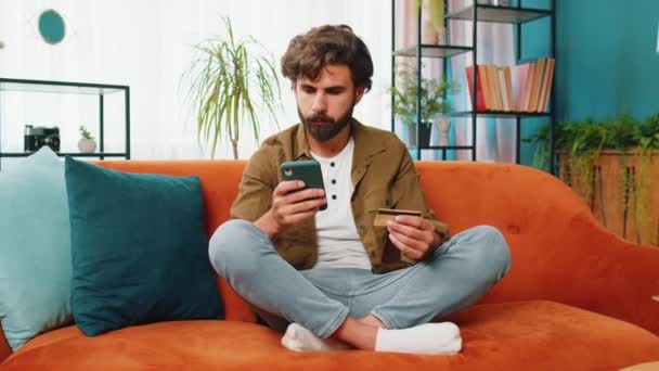 Közel-keleti férfi használja a hitelkártya és az okostelefon, miközben pénzt utalnak, vásárol online vásárlás készpénz nélküli rendelés élelmiszer-szállítás otthon lakás beltéri. Boldog fiatal srác ül a kanapén - Felvétel, videó