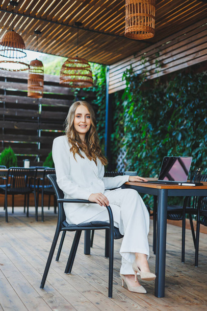 Een mooie vrouw in een wit klassiek pak werkt met een laptop op een open terras in een café. Een vrouw in stijlvolle zakelijke kleding werkt op afstand op een laptop in een zomercafé - Foto, afbeelding