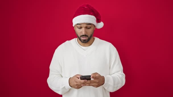 Afričan Američan muž na sobě vánoční klobouk ukazuje na smartphone přes izolované červené pozadí - Záběry, video