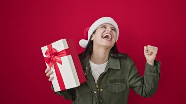 Genç, güzel, İspanyol bir kadın elinde Noel hediyesi tutarak izole edilmiş kırmızı arka planda dans ediyor. - Video, Çekim