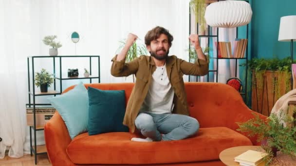 Happy middle eastern man shouting, celebrating success, winning, goal achievement good news, lottery luck, victory, watching TV championship at modern home apartment. Guy in living room sits on couch - Footage, Video