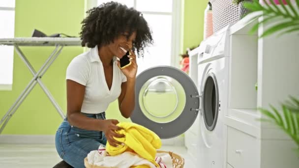 Afroamerykanka rozmawia na smartfonie pranie ubrań uśmiechając się do pralni - Materiał filmowy, wideo
