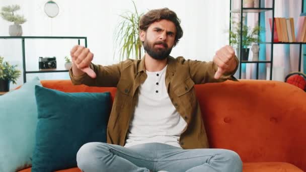 Dislike. Upset unhappy young man showing thumbs down sign gesture, expressing discontent, disapproval dissatisfied bad work at home apartment indoors. Displeased middle eastern guy in room on sofa - Footage, Video