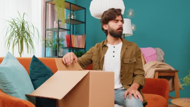 Rozzlobený nespokojený nakupující muž vybalovat balíček pocit rozrušený a zmatený s chybným doručením z internetového obchodu, špatná kvalita rozbitý nákup doma. Arabština chlap uvnitř v pokoji na gauči - Záběry, video