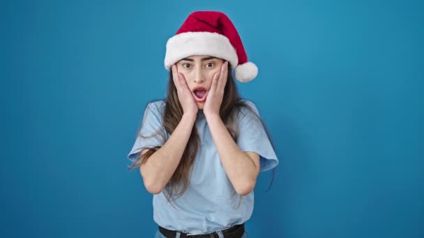 Молодая красивая латиноамериканка удивляет выражение лица в рождественской шляпе на изолированном синем фоне - Кадры, видео
