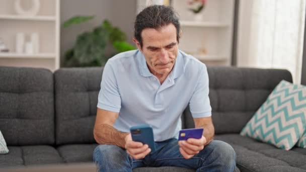 Мужчина среднего возраста делает покупки со смартфоном и кредитной картой, сидя дома на диване - Кадры, видео