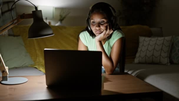 Laptop ve kulaklık kullanan genç latin kadın koltukta oturmuş evde esniyor. - Video, Çekim