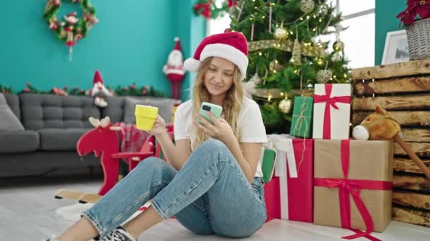 Mujer rubia joven usando smartphone bebiendo café celebrando la Navidad en casa - Imágenes, Vídeo