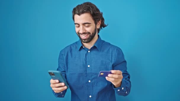 Fiatal spanyol férfi használ okostelefon és hitelkártya nézett ideges elszigetelt kék háttér - Felvétel, videó