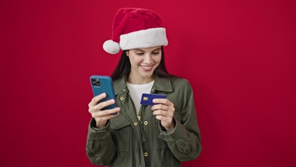 Νέα όμορφη ισπανόφωνη γυναίκα ψώνια με smartphone και πιστωτική κάρτα φορώντας καπέλο Χριστουγέννων πάνω από απομονωμένο κόκκινο φόντο - Πλάνα, βίντεο