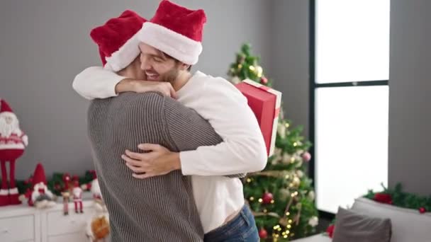 Dwóch mężczyzn świętuje Boże Narodzenie trzymając prezent przytulając się w domu - Materiał filmowy, wideo