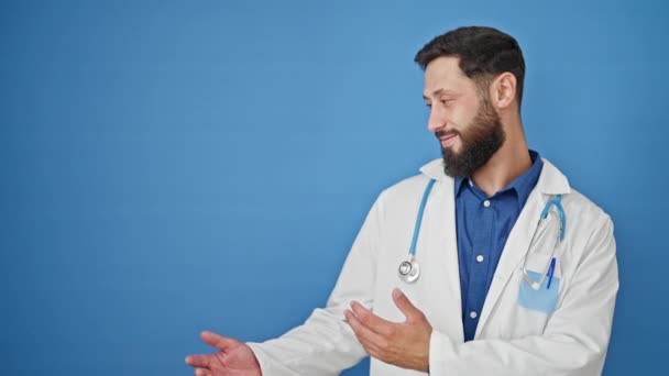 Junger hispanischer Arzt lächelt selbstbewusst und präsentiert sich vor isoliertem blauen Hintergrund - Filmmaterial, Video