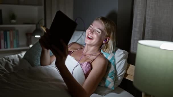Junge blonde Frau sieht sich Film an und lacht viel im Schlafzimmer - Filmmaterial, Video