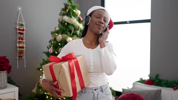 African american woman holding gift by christmas tree dancing at home - Footage, Video