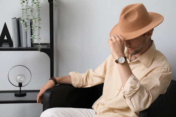 Elegante joven con reloj de pulsera y sombrero sentado en el sillón en la habitación - Foto, imagen